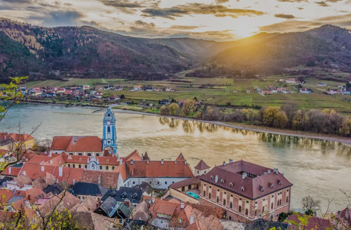 View of Dürnstein and the blue church tower overlooking the Danube River in Austria’s Wachau Valley at sunset, surrounded by vineyards and rolling hills — a highlight of Danube River cruises.